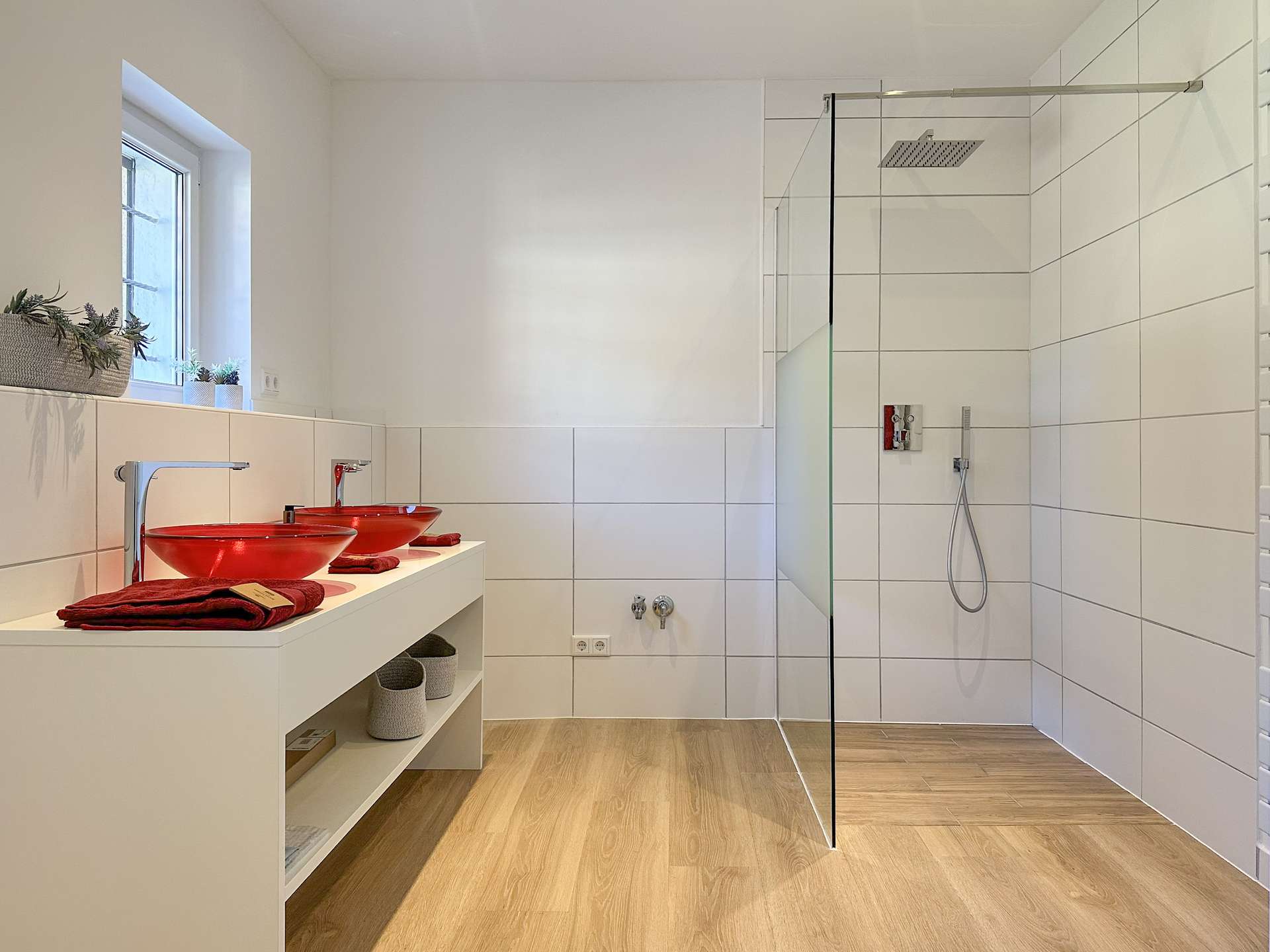 Modern bathroom with white tiles, wood-look floor, and a glass shower enclosure; red vessel sinks on a white vanity. Gently lit by a window.",