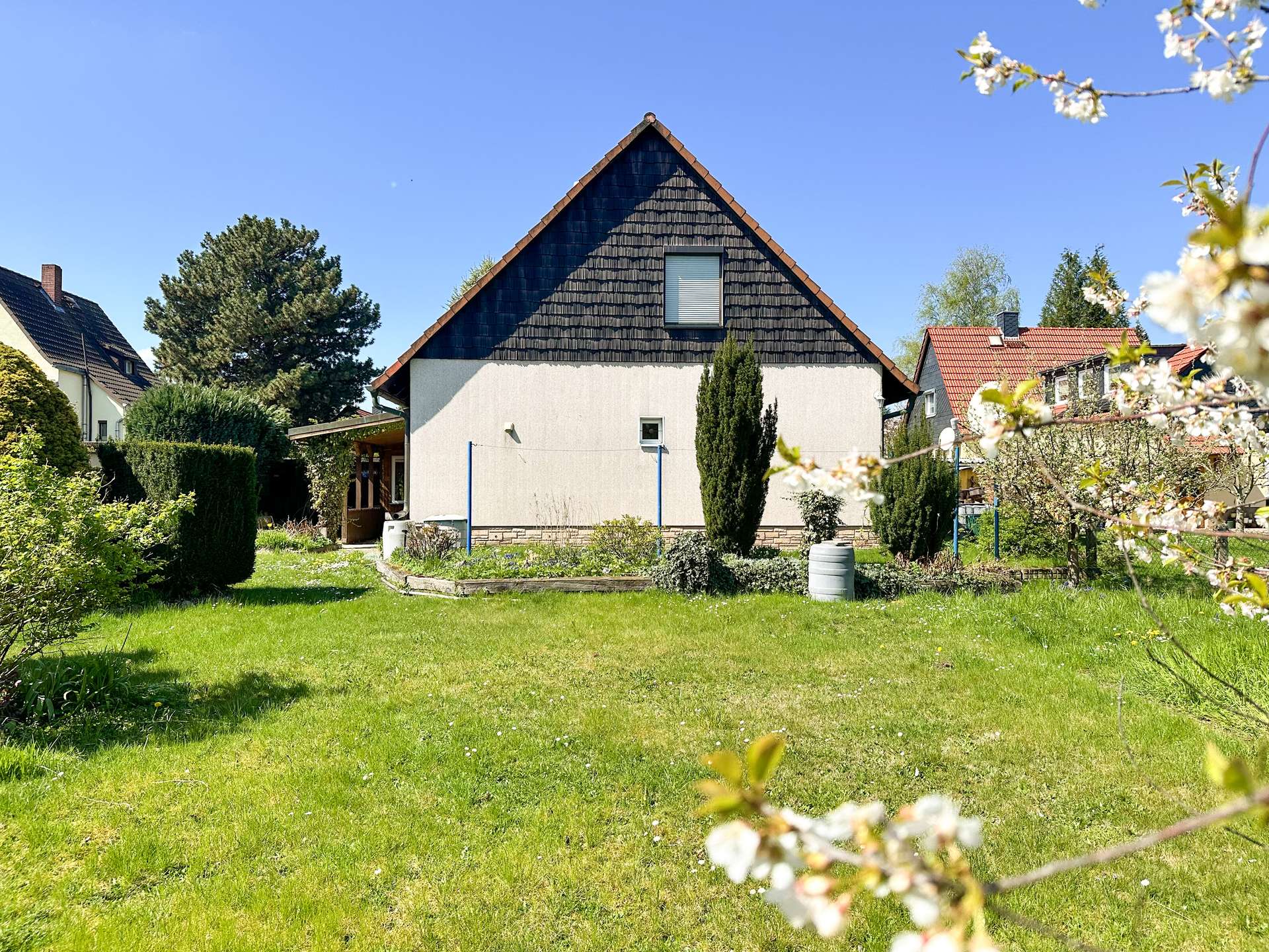 Two-story house with a steep triangular roof in a sunny garden, foreground flowering branches, blue sky above.