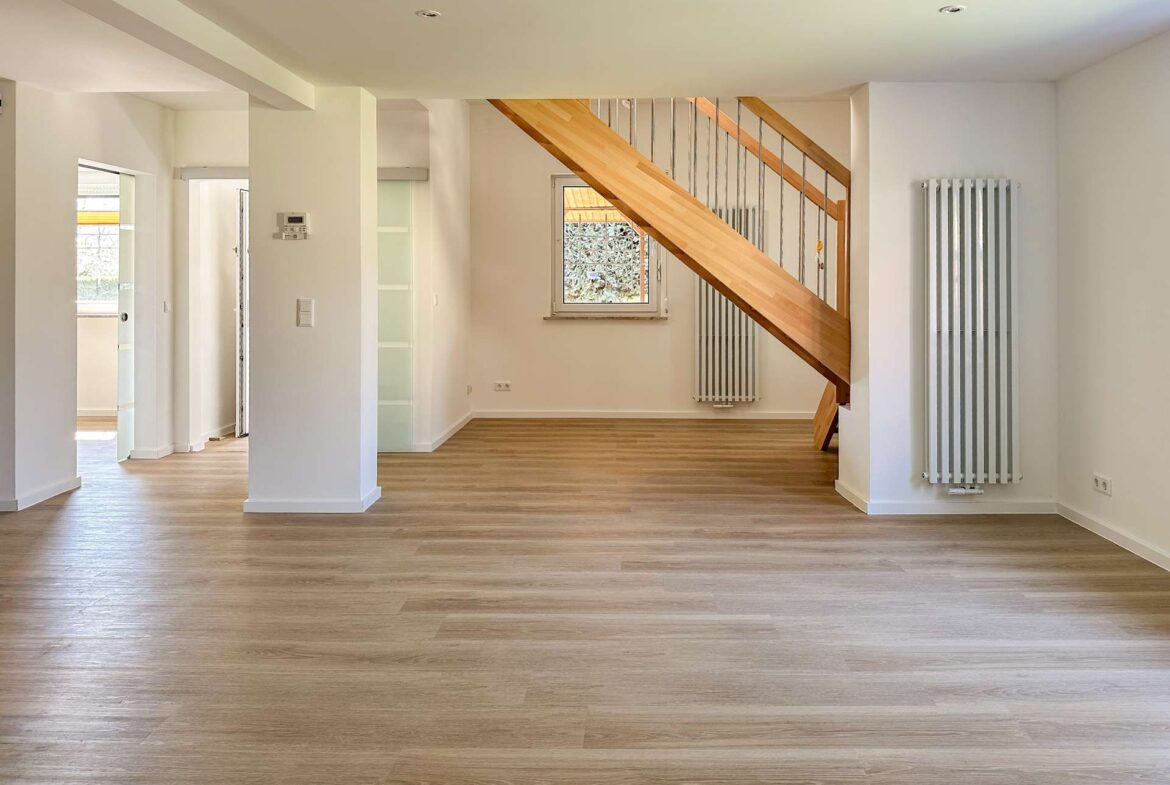 Bright open-plan living area with light wood flooring, a wooden staircase with metal balusters, and white walls.