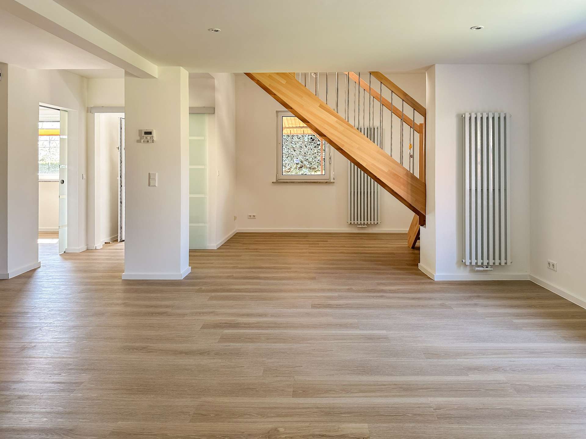 Bright open-plan living area with light wood flooring, a wooden staircase with metal balusters, and white walls.