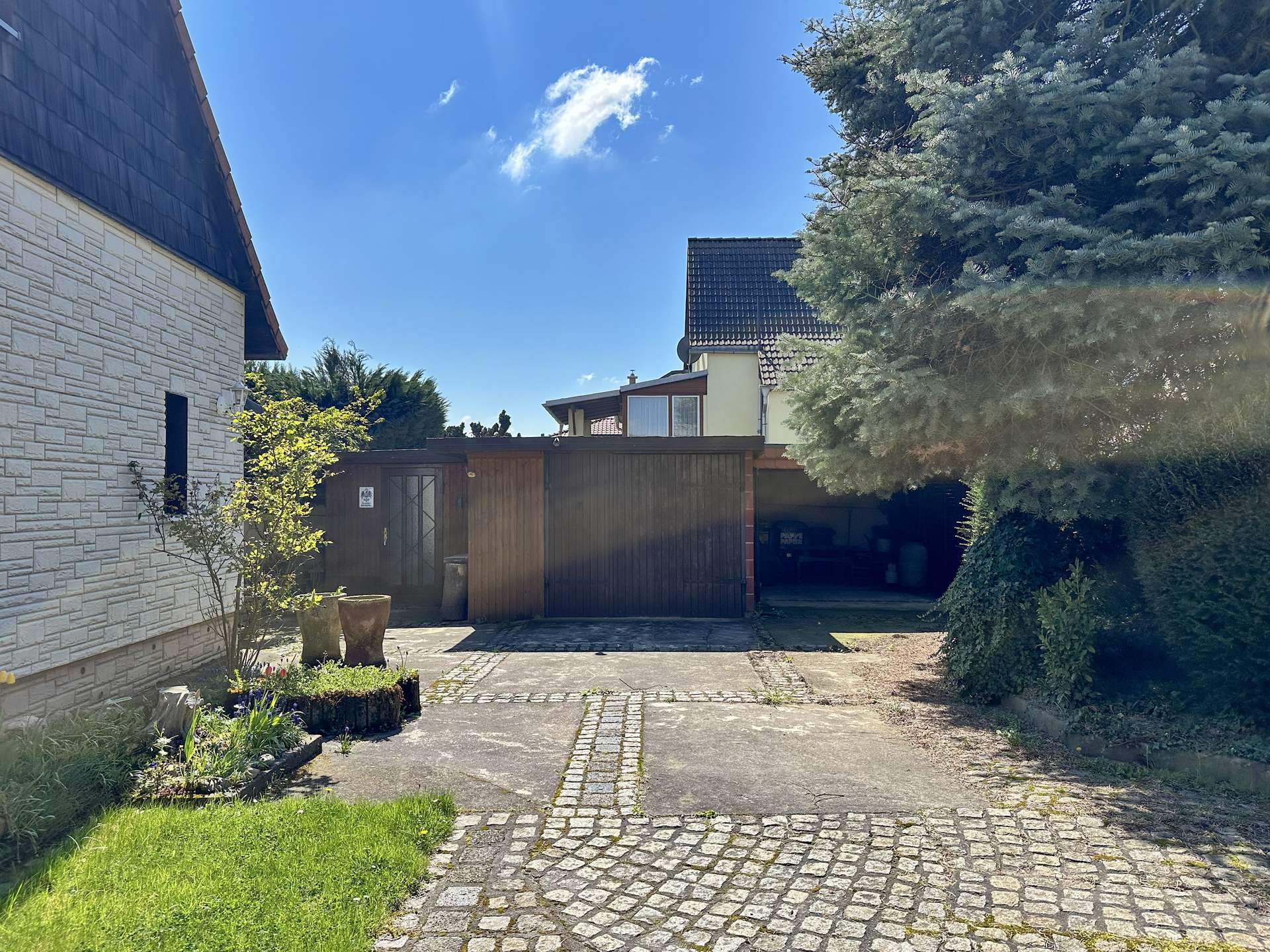 Sunny residential driveway with a stone-sided house on the left, a wooden gate in the center, and a large evergreen tree on the right under a blue sky.