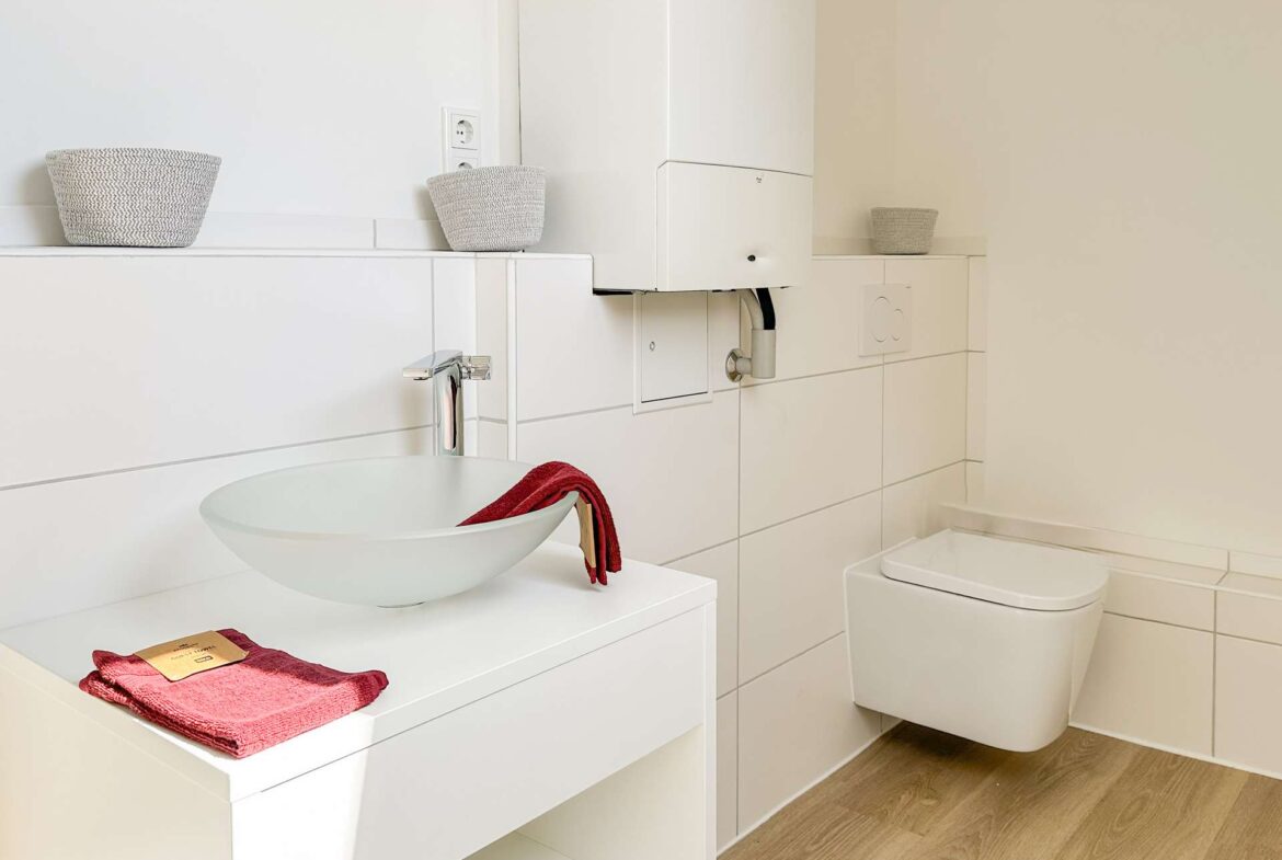Modern white bathroom with a round vessel sink on a white vanity, red towel, wicker baskets, and a wall-mounted boiler behind the sink.