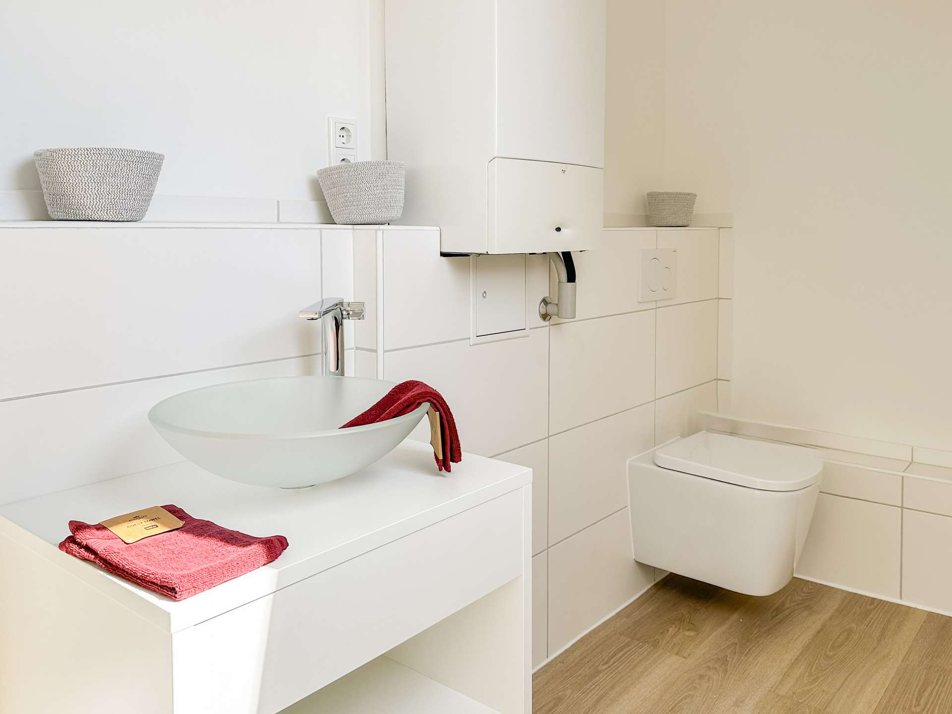 Modern white bathroom with a round vessel sink on a white vanity, red towel, wicker baskets, and a wall-mounted boiler behind the sink.