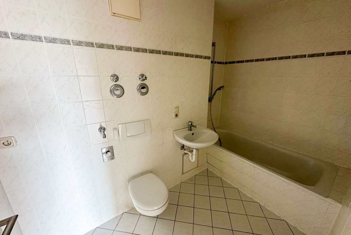 Small bathroom with a toilet, wall sink, and bathtub/shower combo amid white tiled walls and floor.