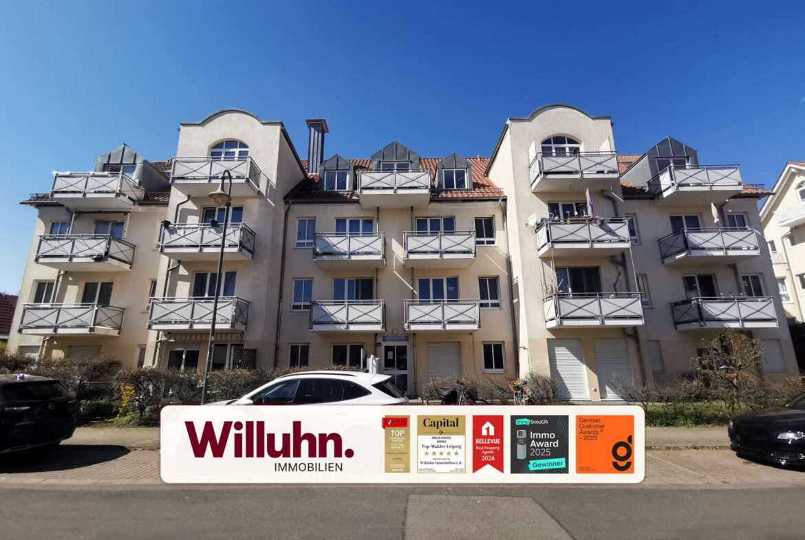 Cream-colored apartment building with numerous balconies and a large Willuhn Immobilien banner at street level against a clear blue sky.