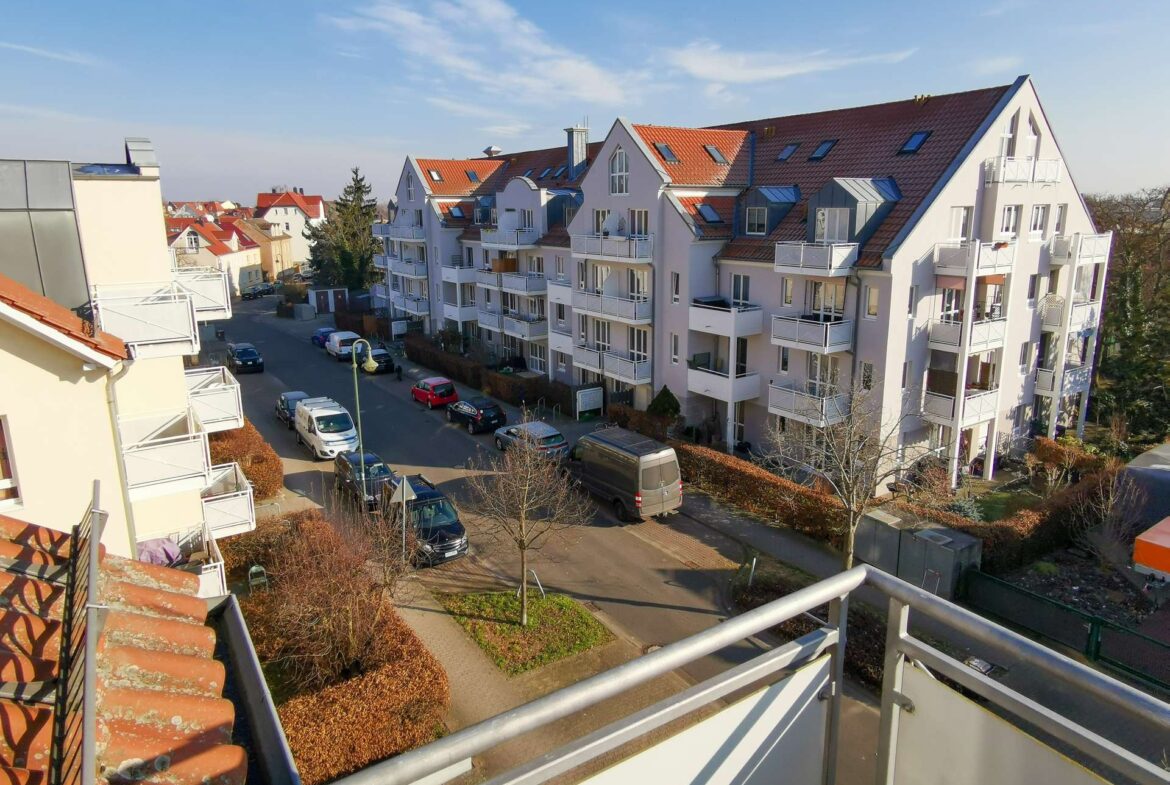 Row of white apartment buildings with balconies and red-tiled roofs along a sunny street, cars parked below.