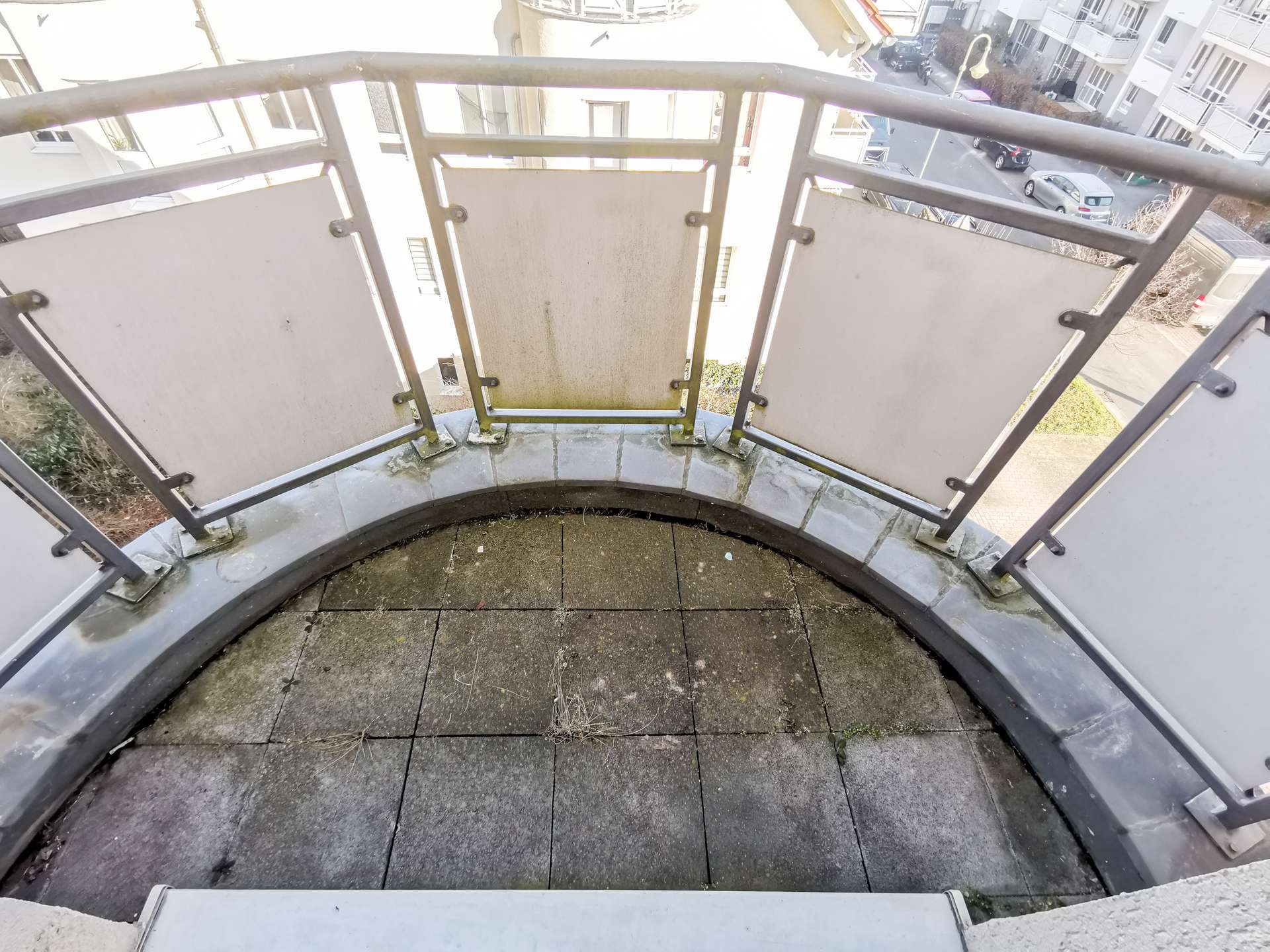 Curved apartment balcony with metal railing and dirty panels, looking down at a street with parked cars and surrounding white buildings.