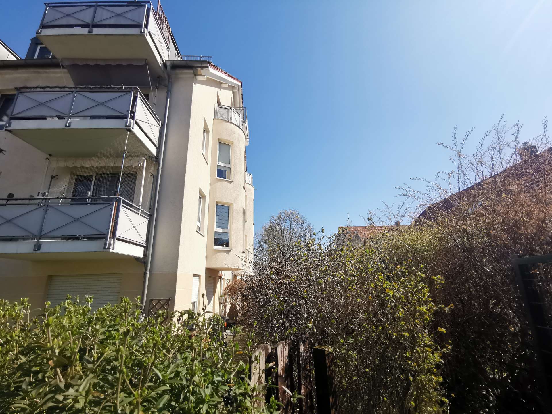 Apartment building with protruding balconies on the left, clear blue sky above and bare shrubs in the foreground.