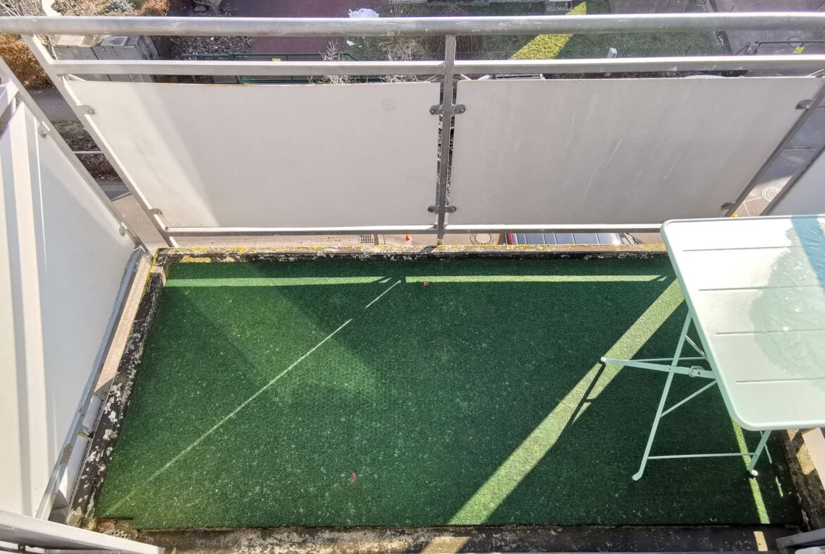 High-angle view of a small balcony with green artificial turf, metal railing, and a pale green folding chair beside a glass table; street and neighboring buildings are visible beyond the railing.