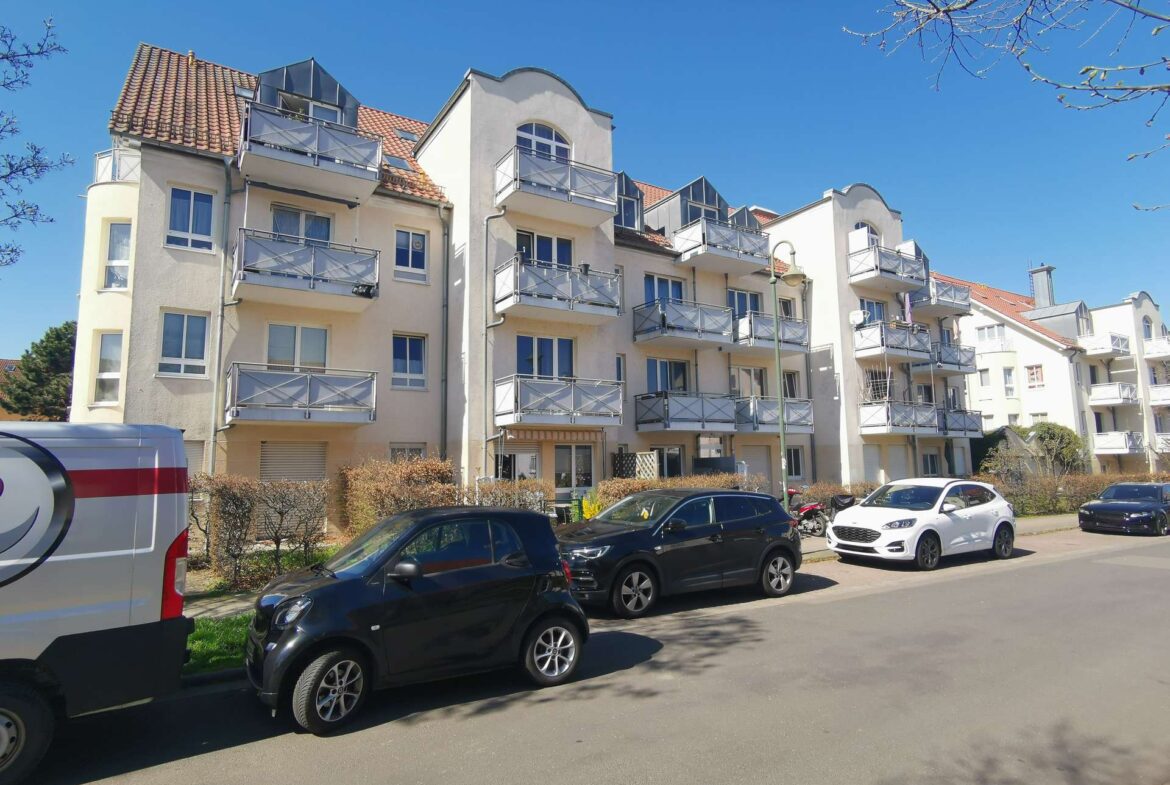 Row of beige apartment buildings with balconies under a clear blue sky; cars parked along the street in front.