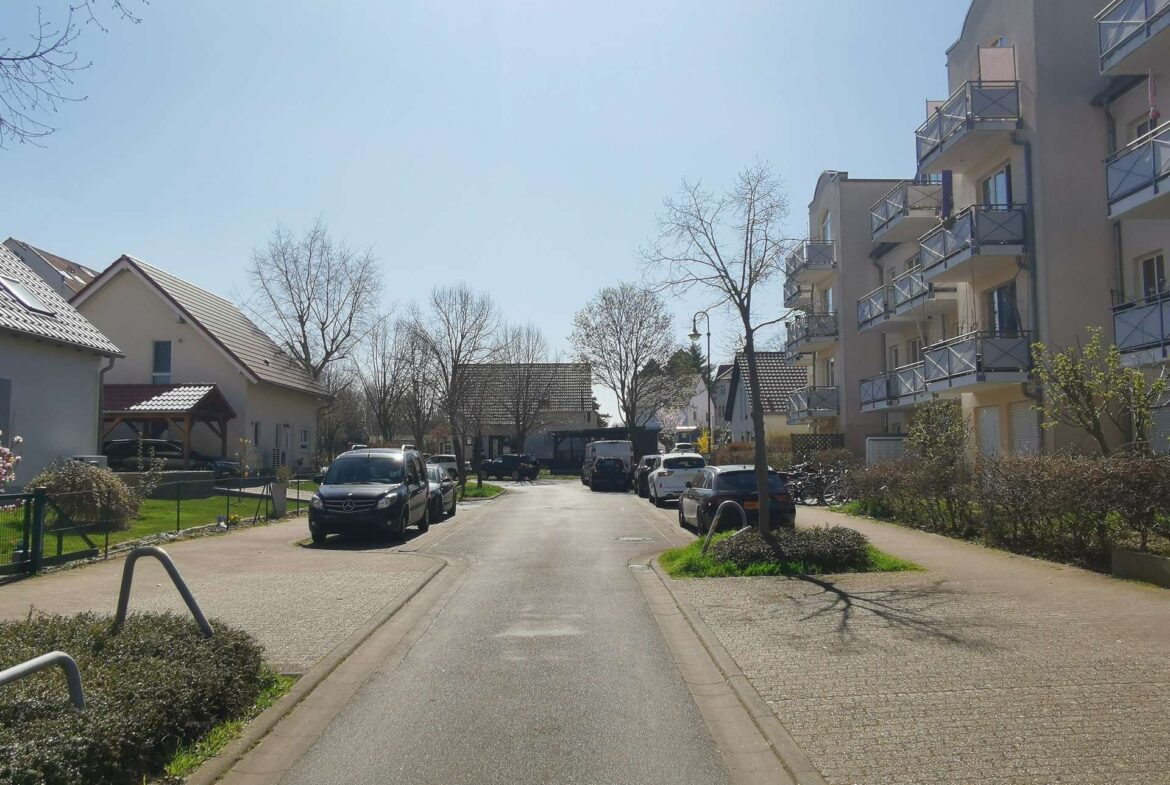 Residential street lined with parked cars and a tall apartment building on the right under a clear blue sky, daytime
