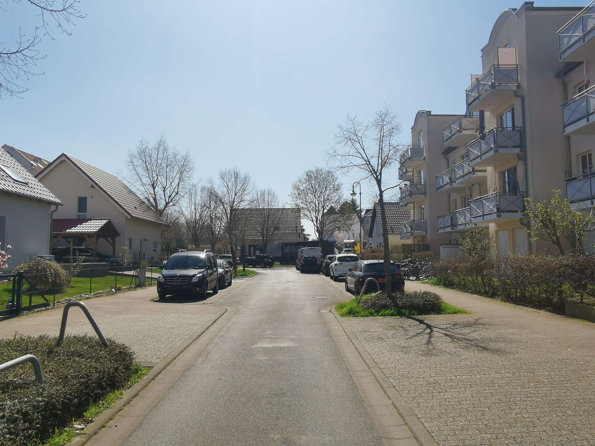 Residential street lined with parked cars and a tall apartment building on the right under a clear blue sky, daytime