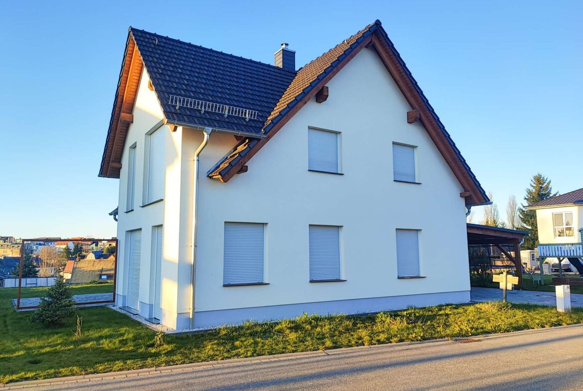White two-story house with a steep dark tiled roof, white exterior walls, and shuttered windows on a sunny street.