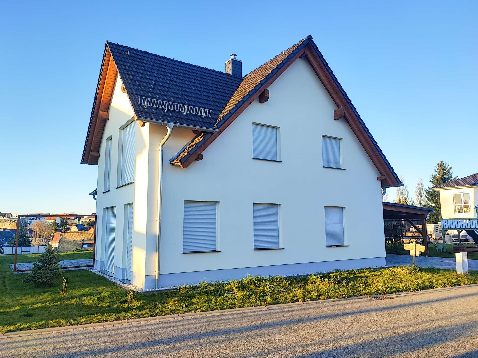 White two-story house with a steep dark tiled roof, white exterior walls, and shuttered windows on a sunny street.