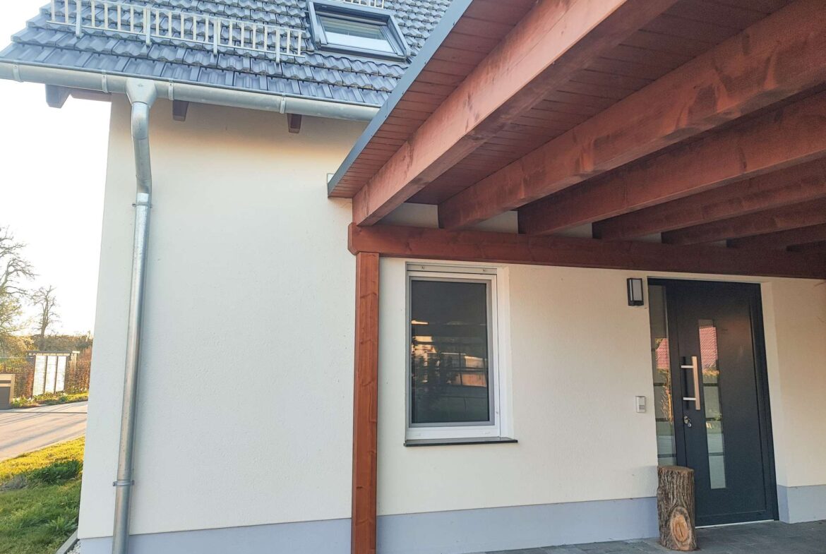 Front entrance of a modern house with a dark gray door under a wooden porch, white wall, a window to the left, and a log stump by the door; pitched tiled roof with a dormer.