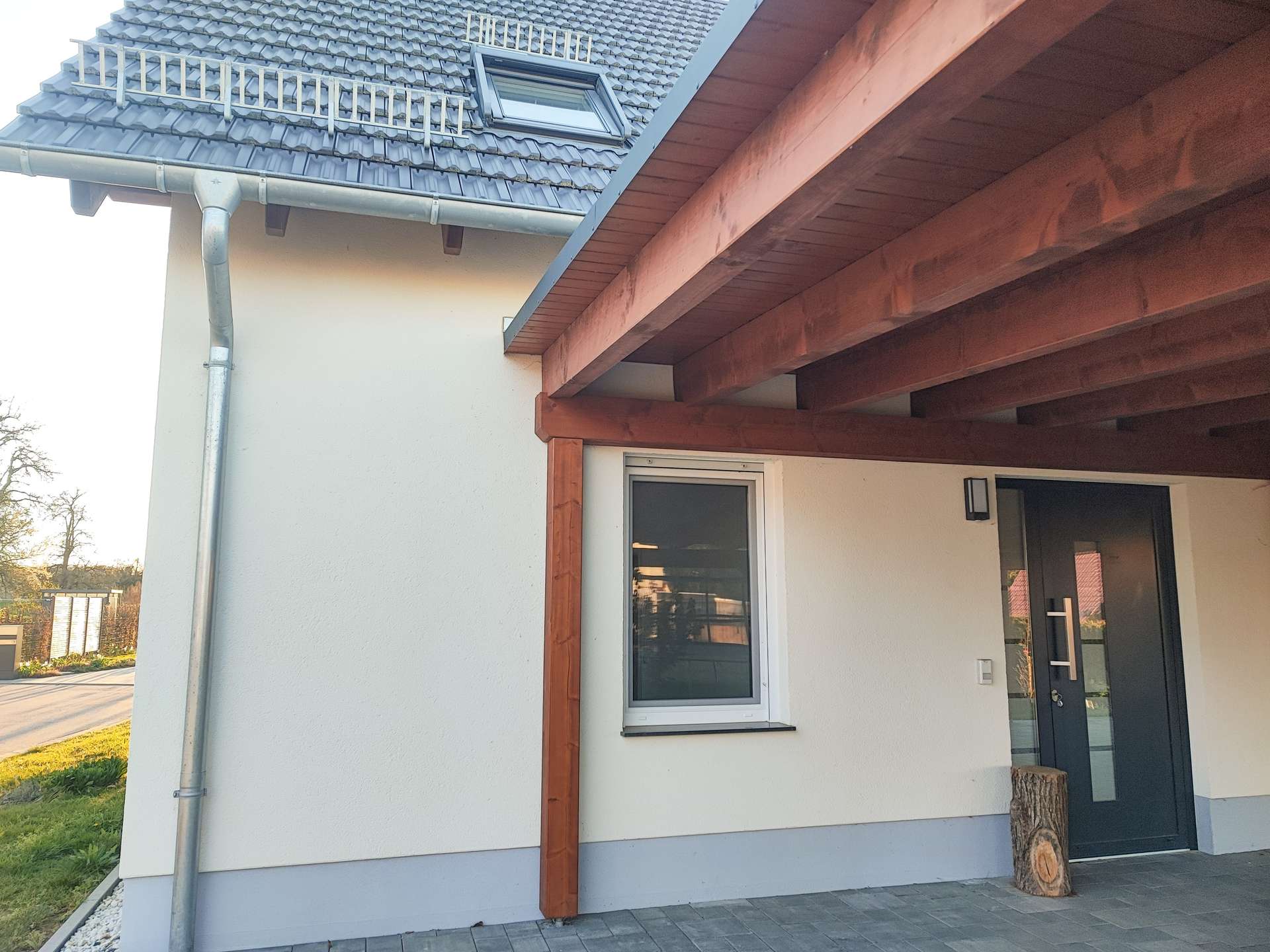 Front entrance of a modern house with a dark gray door under a wooden porch, white wall, a window to the left, and a log stump by the door; pitched tiled roof with a dormer.