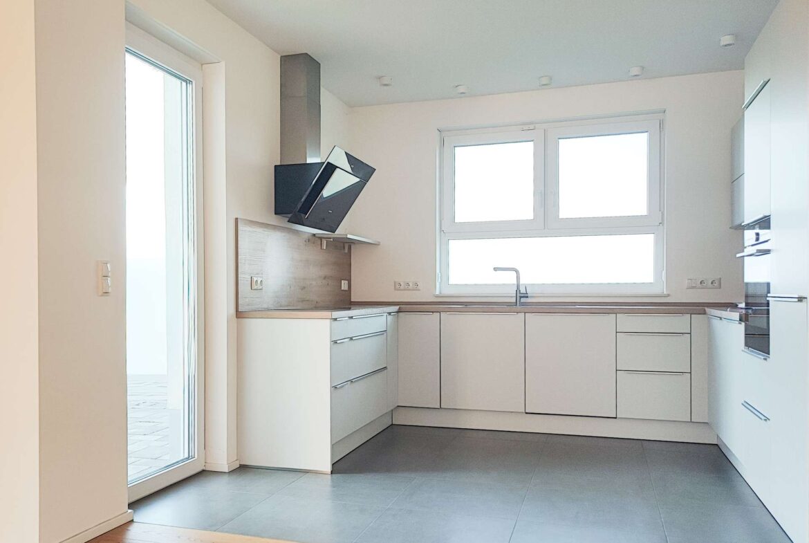 Modern L-shaped white kitchen with wooden countertop, gray floor tiles, and large window over the sink; built-in appliances on the right and a tilted range hood above the stove area.