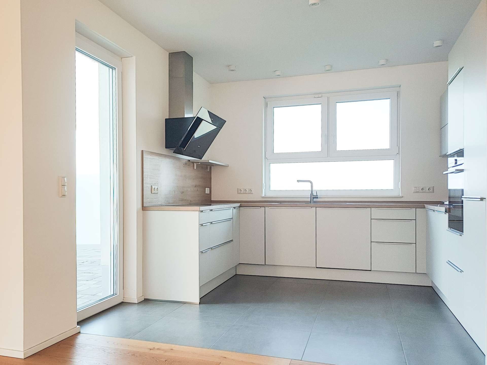 Modern L-shaped white kitchen with wooden countertop, gray floor tiles, and large window over the sink; built-in appliances on the right and a tilted range hood above the stove area.