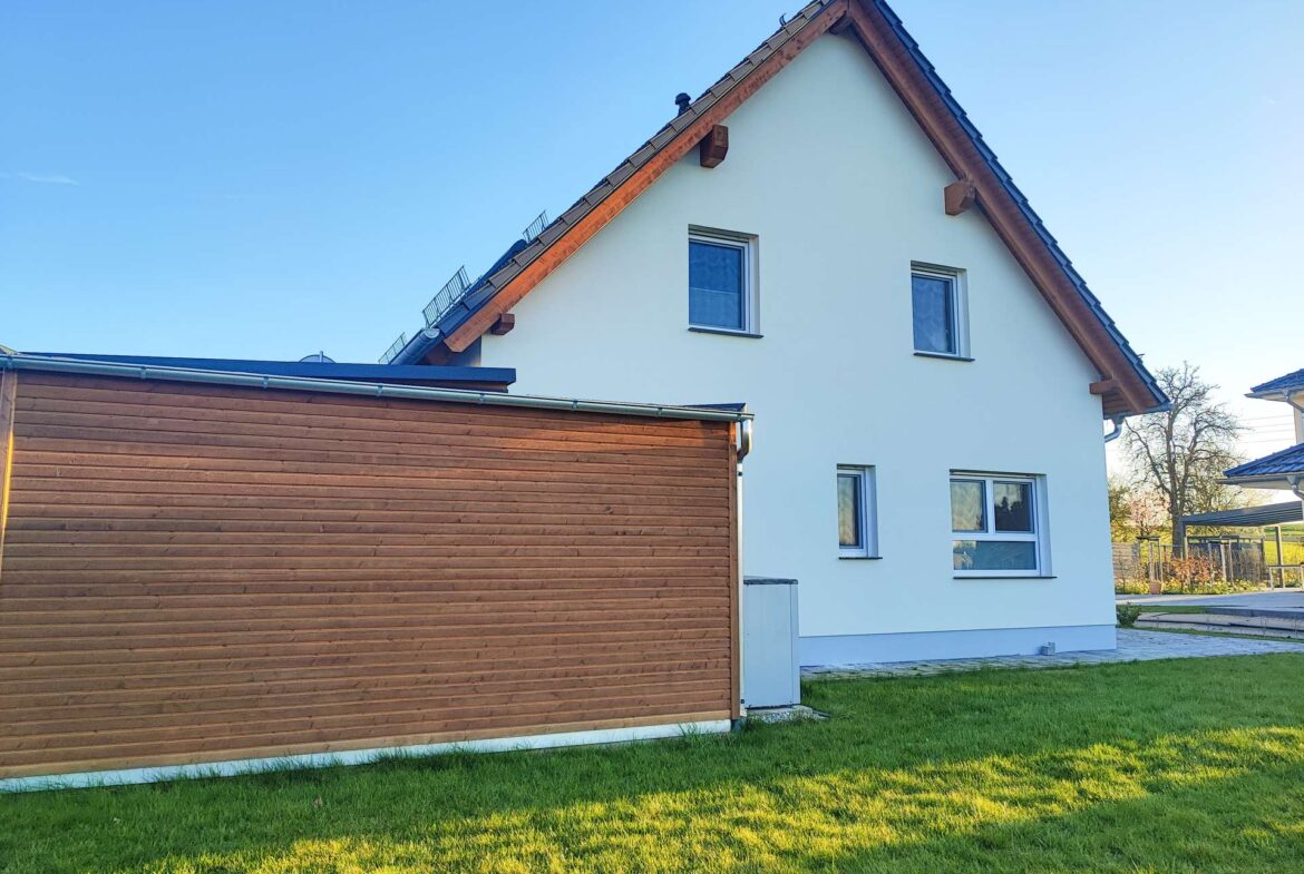 White two-story house with steep pitched roof beside a wooden-clad garage, on a sunny day with a green lawn.