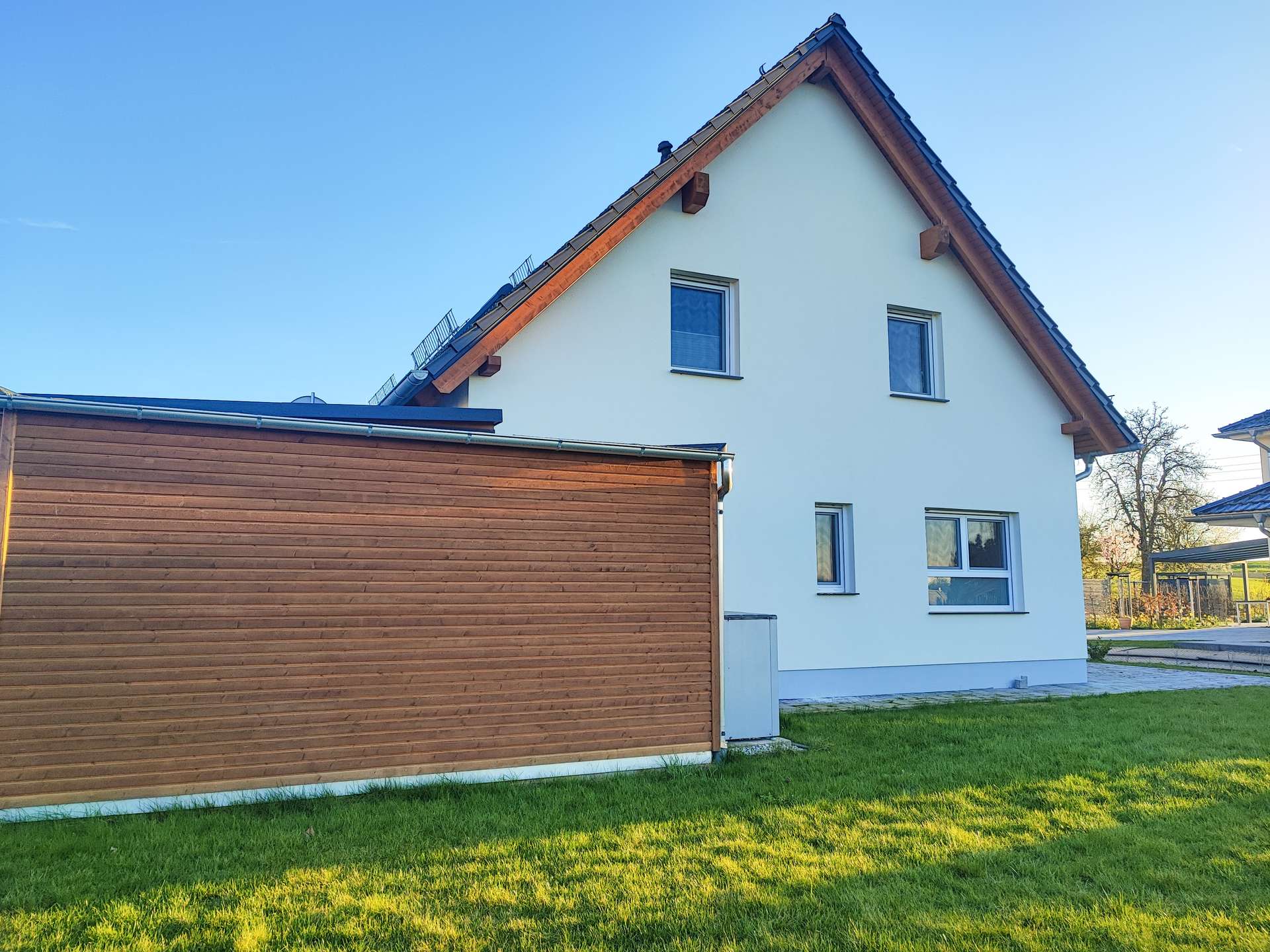 White two-story house with steep pitched roof beside a wooden-clad garage, on a sunny day with a green lawn.