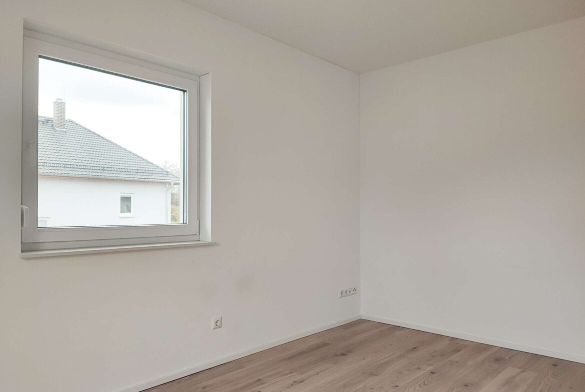 Empty white-walled room with light wood floor and a large window letting in daylight.