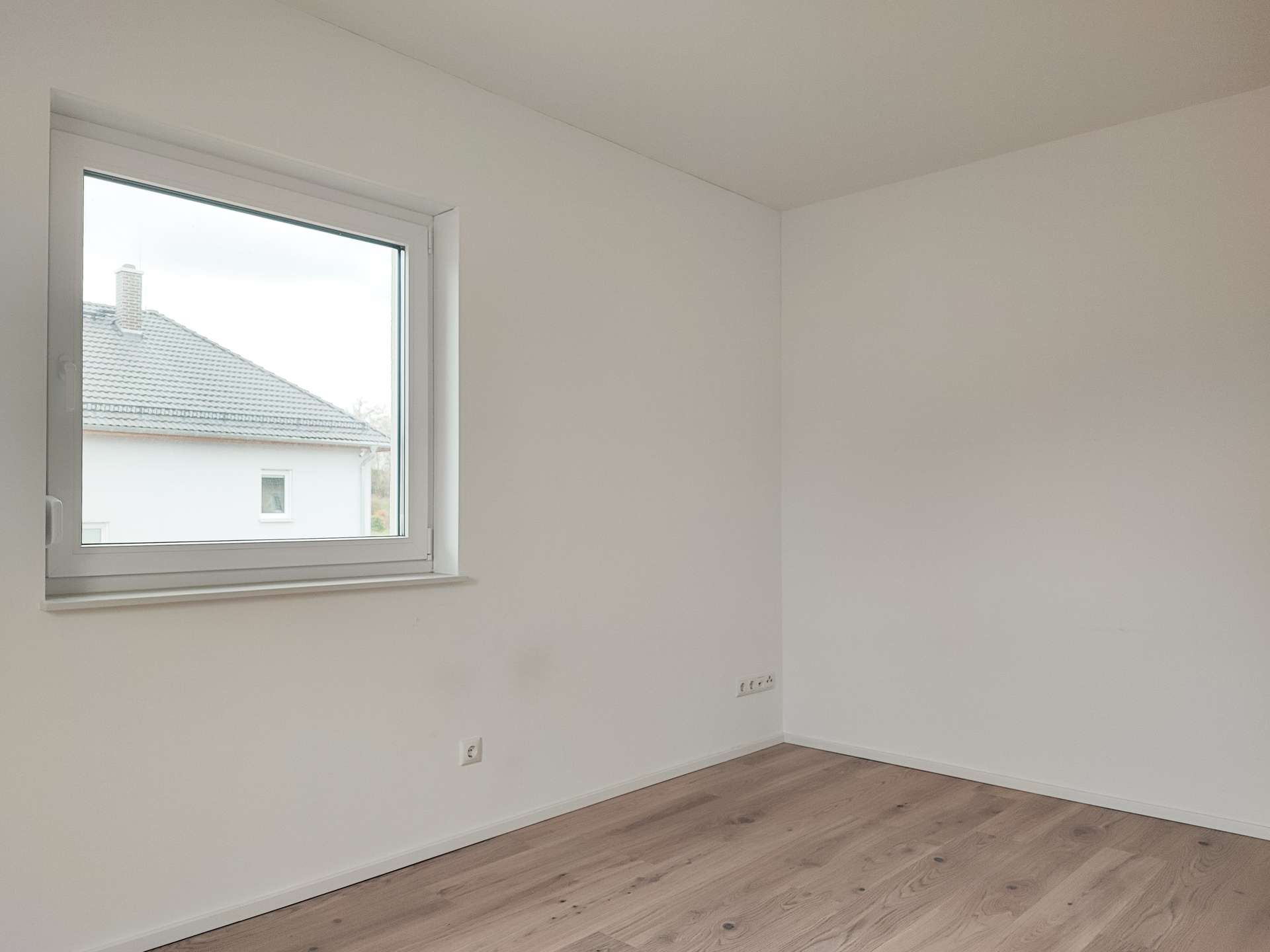 Empty white-walled room with light wood floor and a large window letting in daylight.