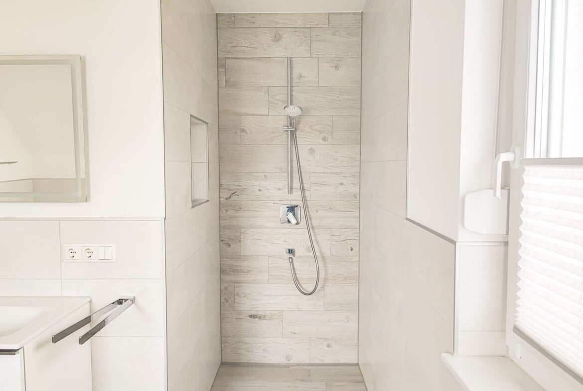 Bright white bathroom with a walk‑in shower featuring light wood‑grain wall tiles and a handheld showerhead, plus a built‑in shower niche and window with blinds on the right.