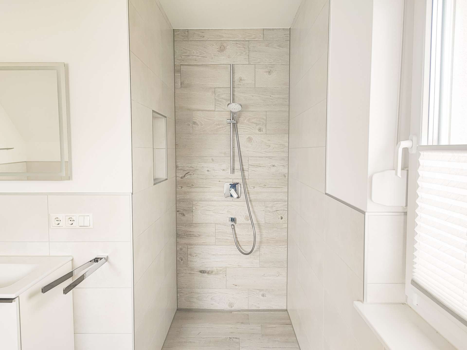 Bright white bathroom with a walk‑in shower featuring light wood‑grain wall tiles and a handheld showerhead, plus a built‑in shower niche and window with blinds on the right.