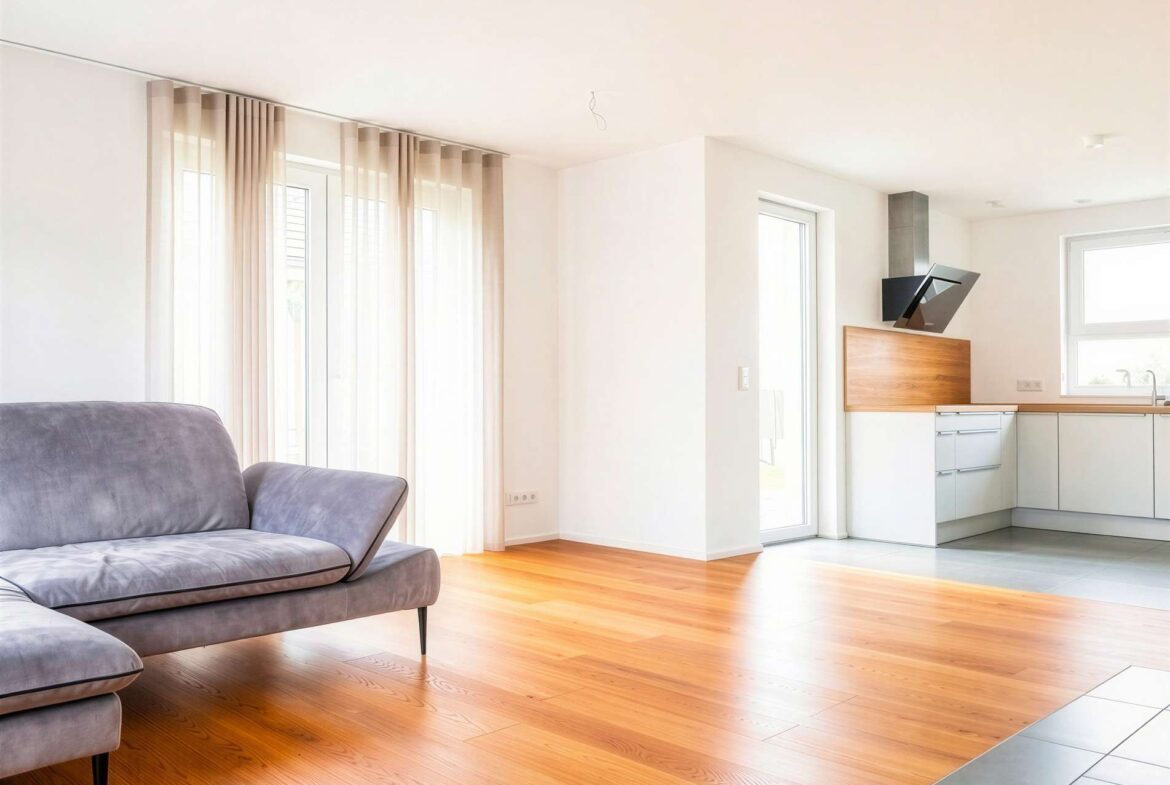 Bright open-plan living area with a gray sofa, warm wood floors, and sheer curtain-covered windows connecting to a modern kitchen.