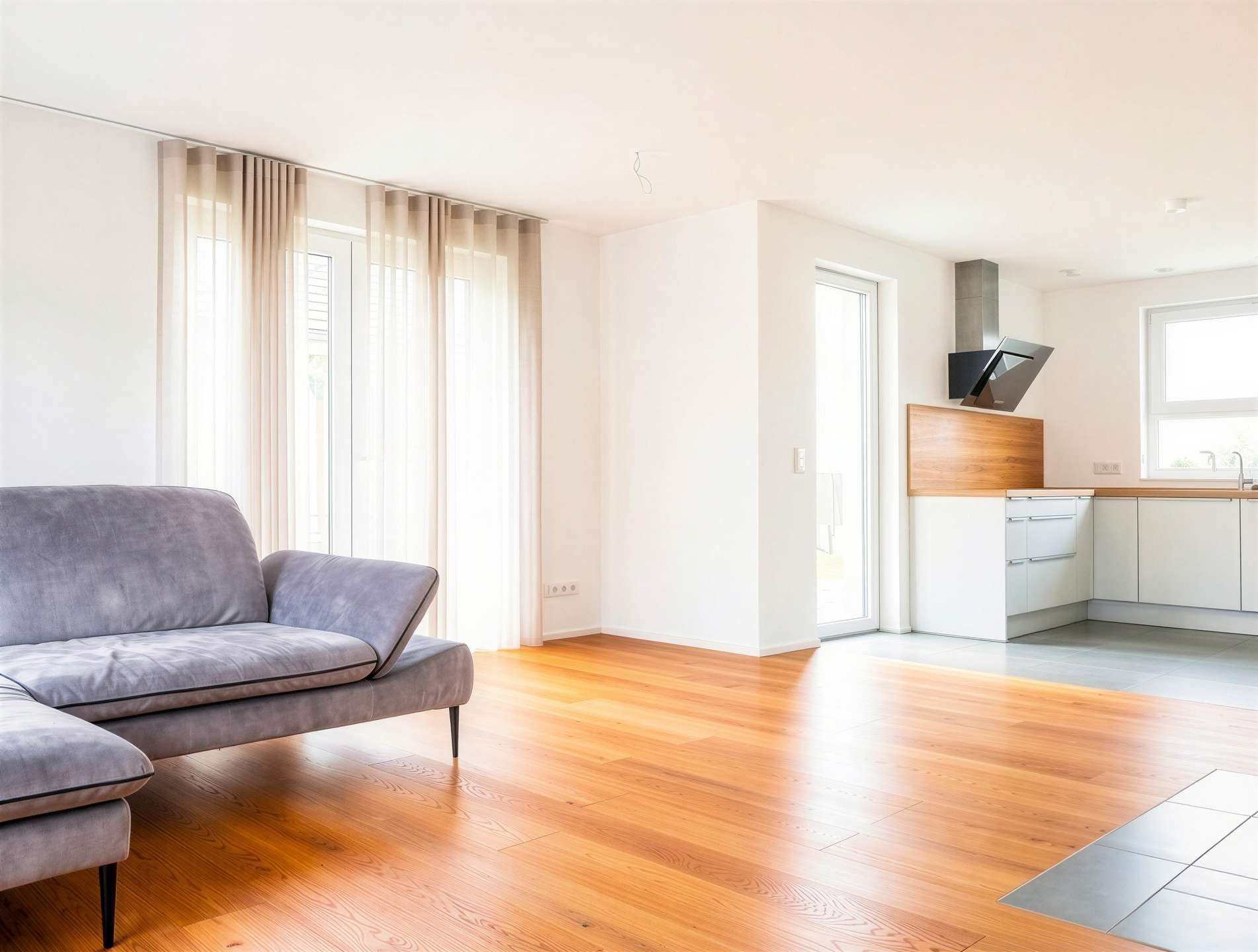 Bright open-plan living area with a gray sofa, warm wood floors, and sheer curtain-covered windows connecting to a modern kitchen.