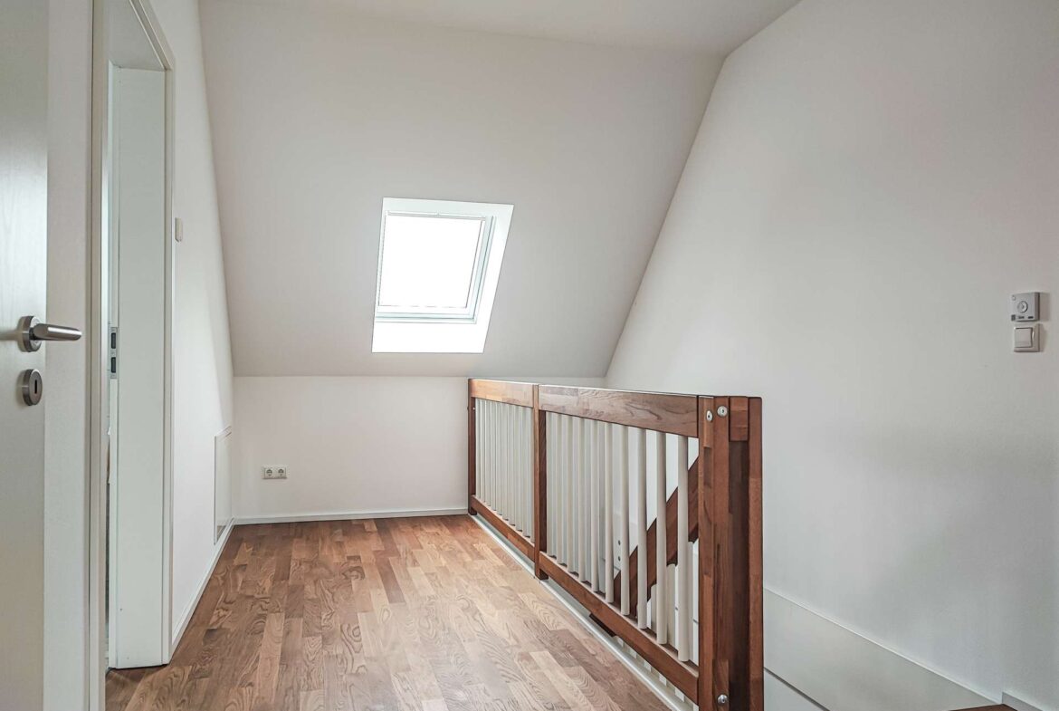 Bright loft landing with a skylight, white walls, and a wooden railing overlooking a staircase on the right.