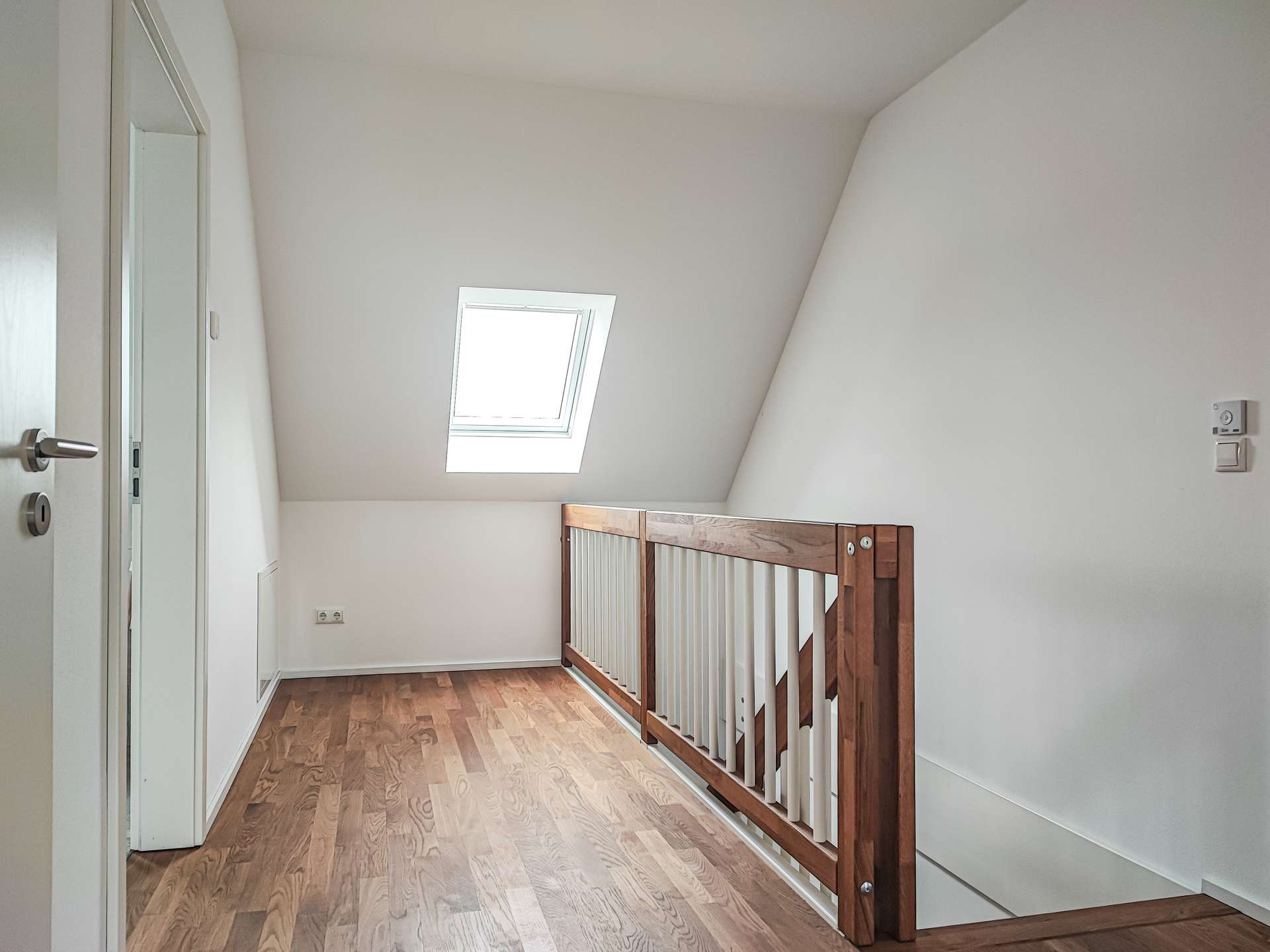 Bright loft landing with a skylight, white walls, and a wooden railing overlooking a staircase on the right.