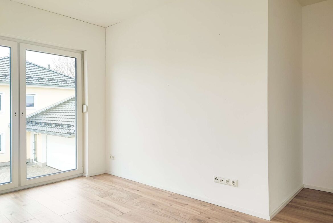 Empty sunlit room with light wood floor and large glass doors opening to an exterior courtyard/yard area.