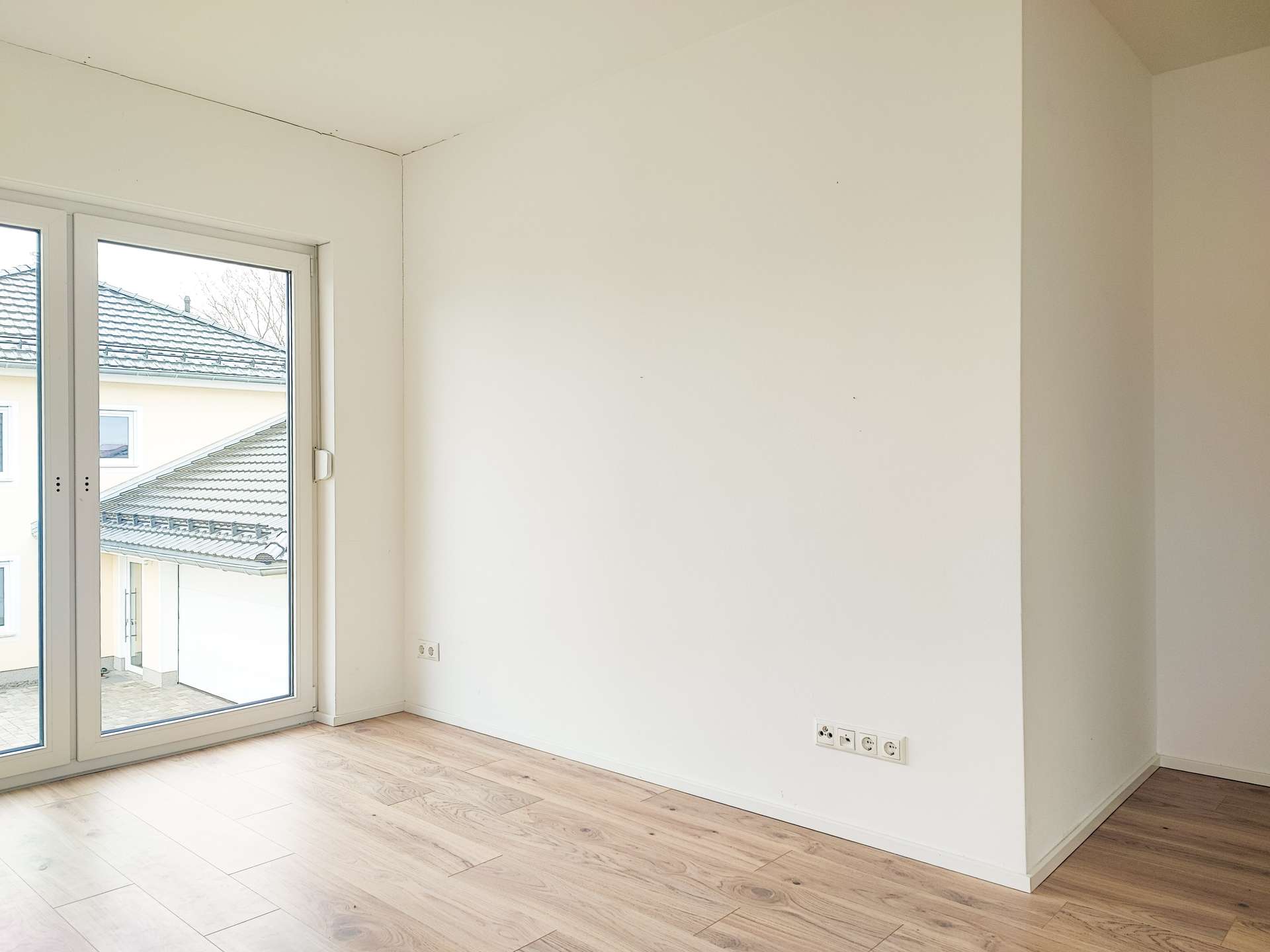 Empty sunlit room with light wood floor and large glass doors opening to an exterior courtyard/yard area.