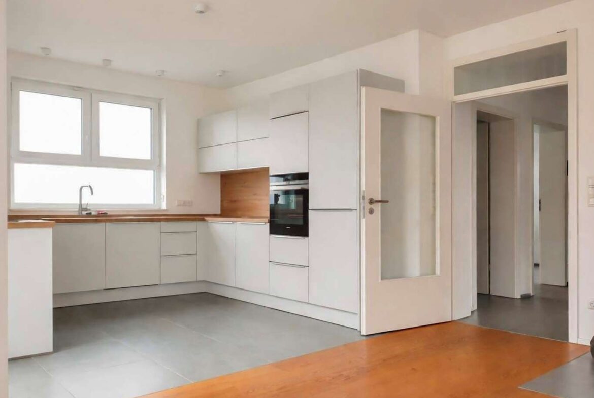 Bright modern kitchen with white cabinets, wooden countertops, and a built-in oven. An open glass-panel door leads to a hallway.