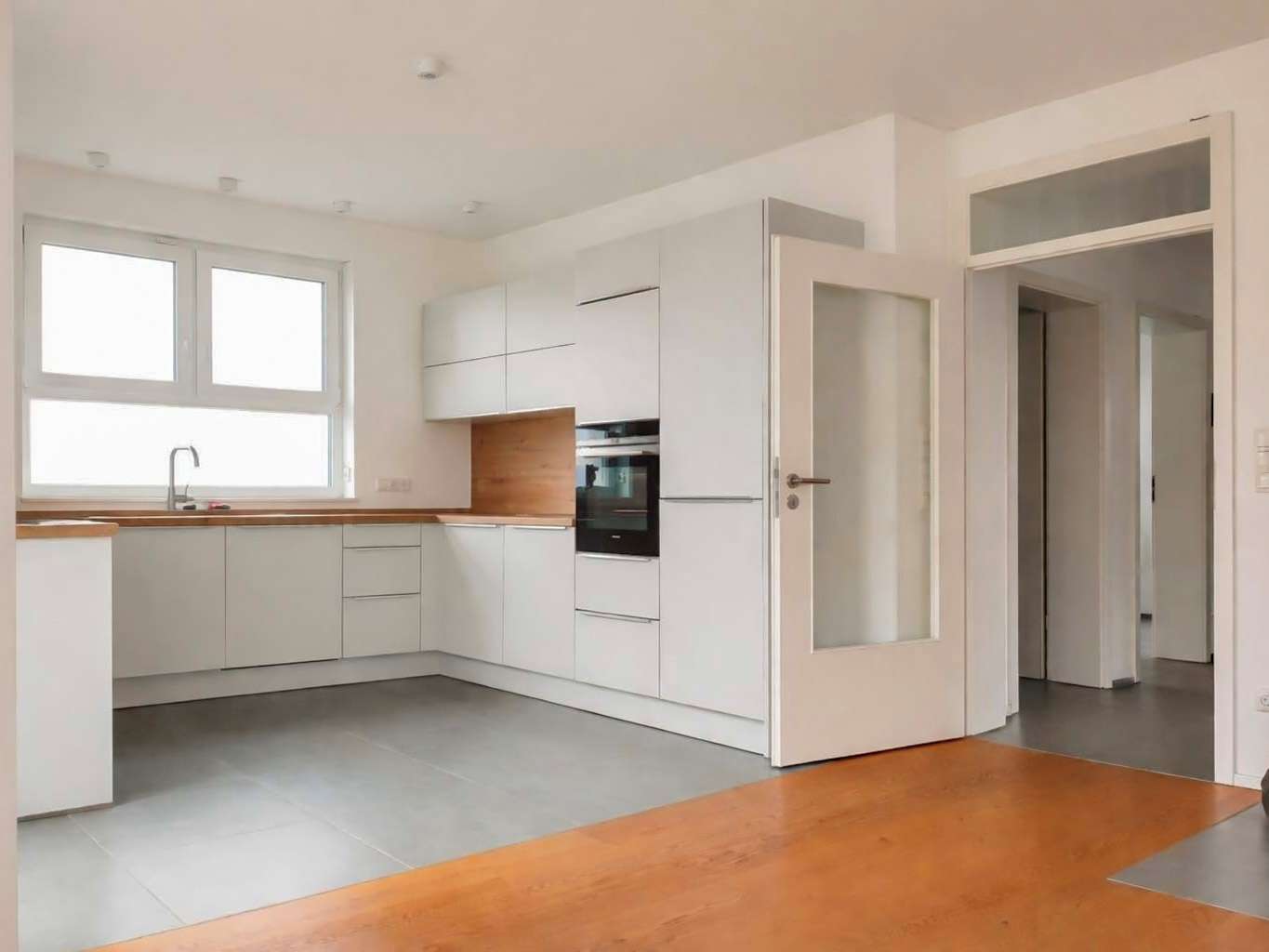 Bright modern kitchen with white cabinets, wooden countertops, and a built-in oven. An open glass-panel door leads to a hallway.