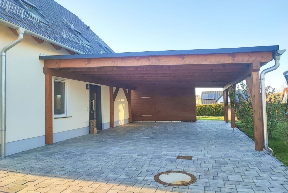 House with a large wooden carport attached to the side, over a gray paver driveway and clear blue sky on a sunny day.