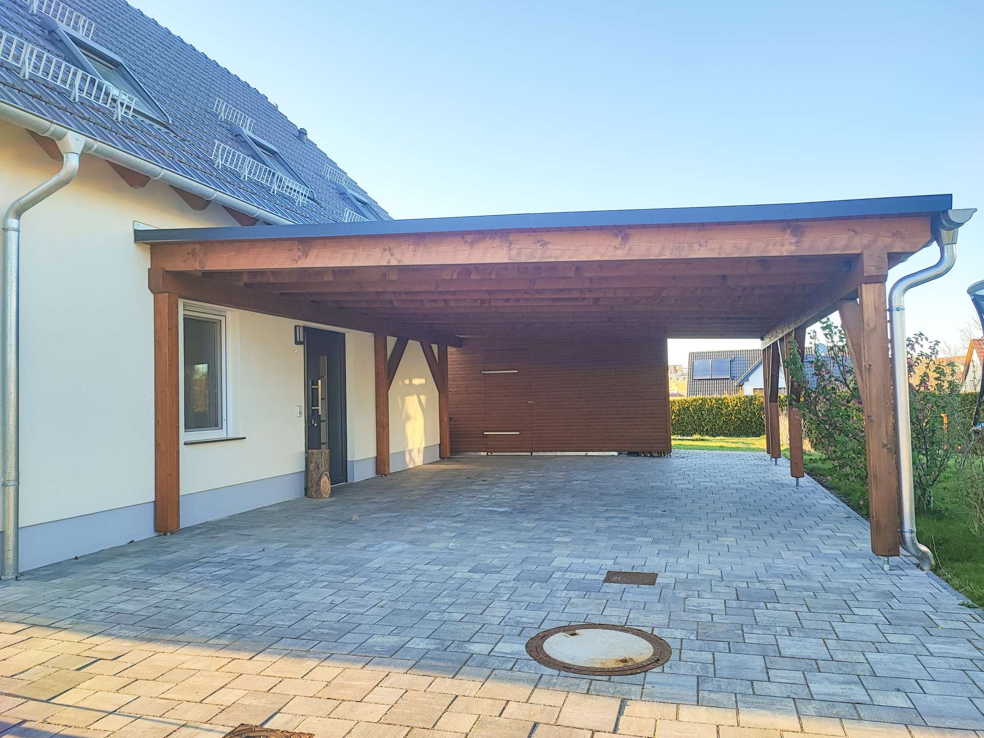 House with a large wooden carport attached to the side, over a gray paver driveway and clear blue sky on a sunny day.