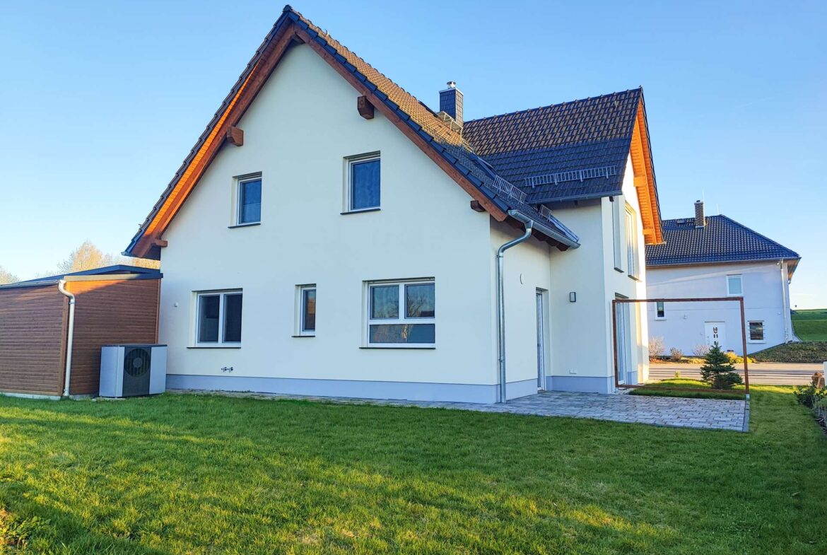 White two-story house with a steep dark tiled roof, wooden eaves, and a well-kept green lawn under a clear blue sky.