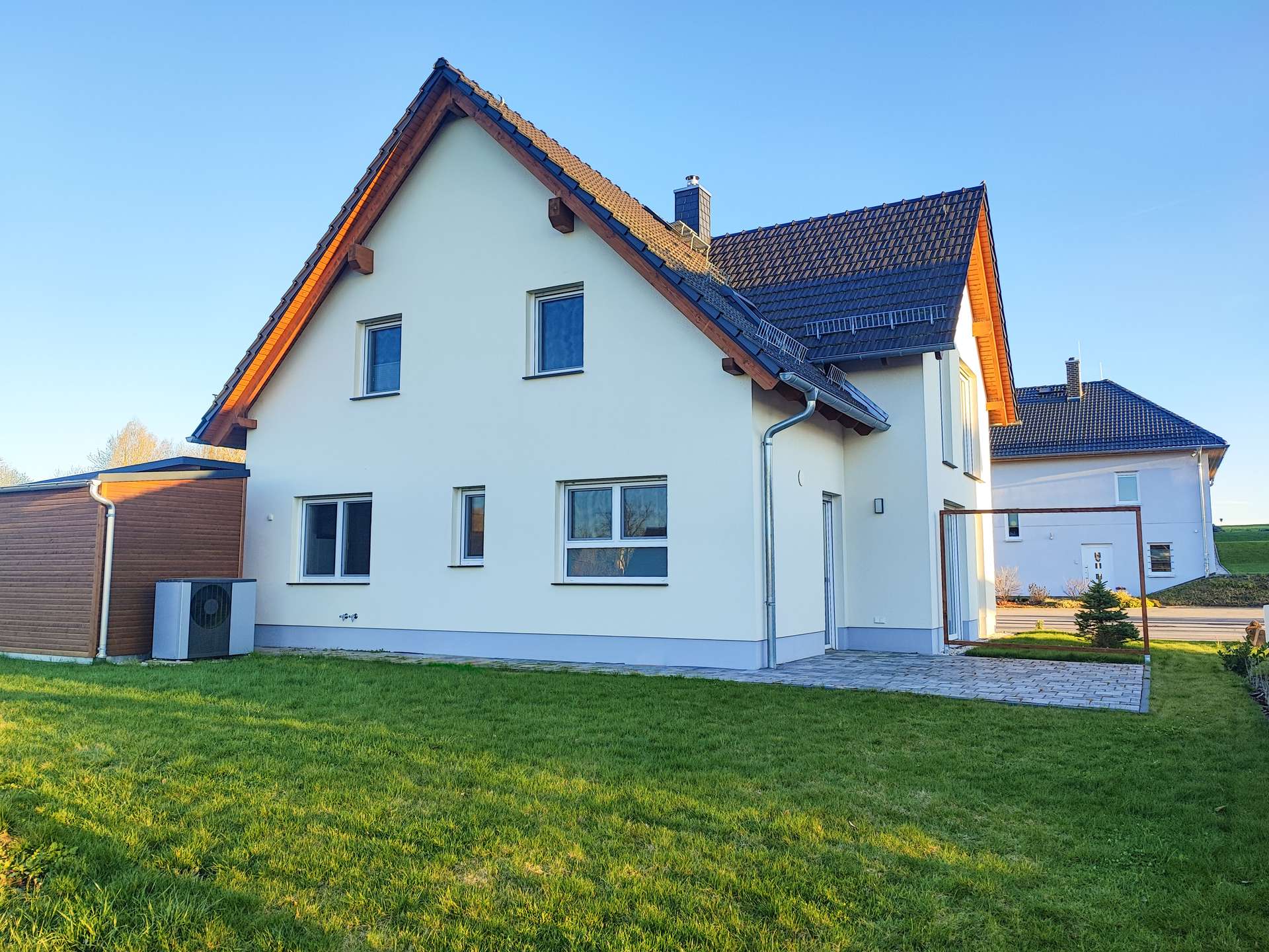 White two-story house with a steep dark tiled roof, wooden eaves, and a well-kept green lawn under a clear blue sky.