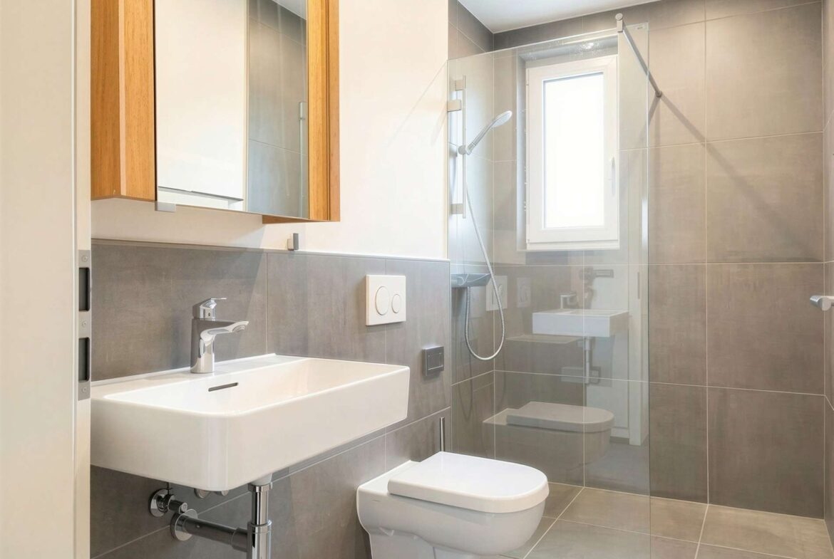 Modern bathroom with white wall-mounted sink, wall-hung toilet, gray tile walls and floor, and a glass shower enclosure.