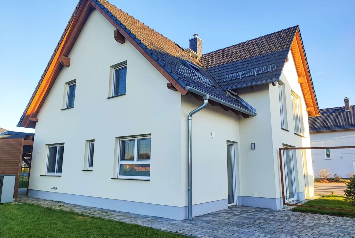 Modern white two-story house with blue trim, steep gabled roof, and a paved patio area in front.