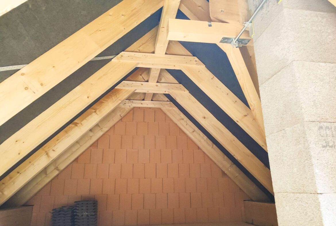 Unfinished attic with exposed wooden roof trusses, brick back wall at the end, and a small stack of roofing tiles on the floor.