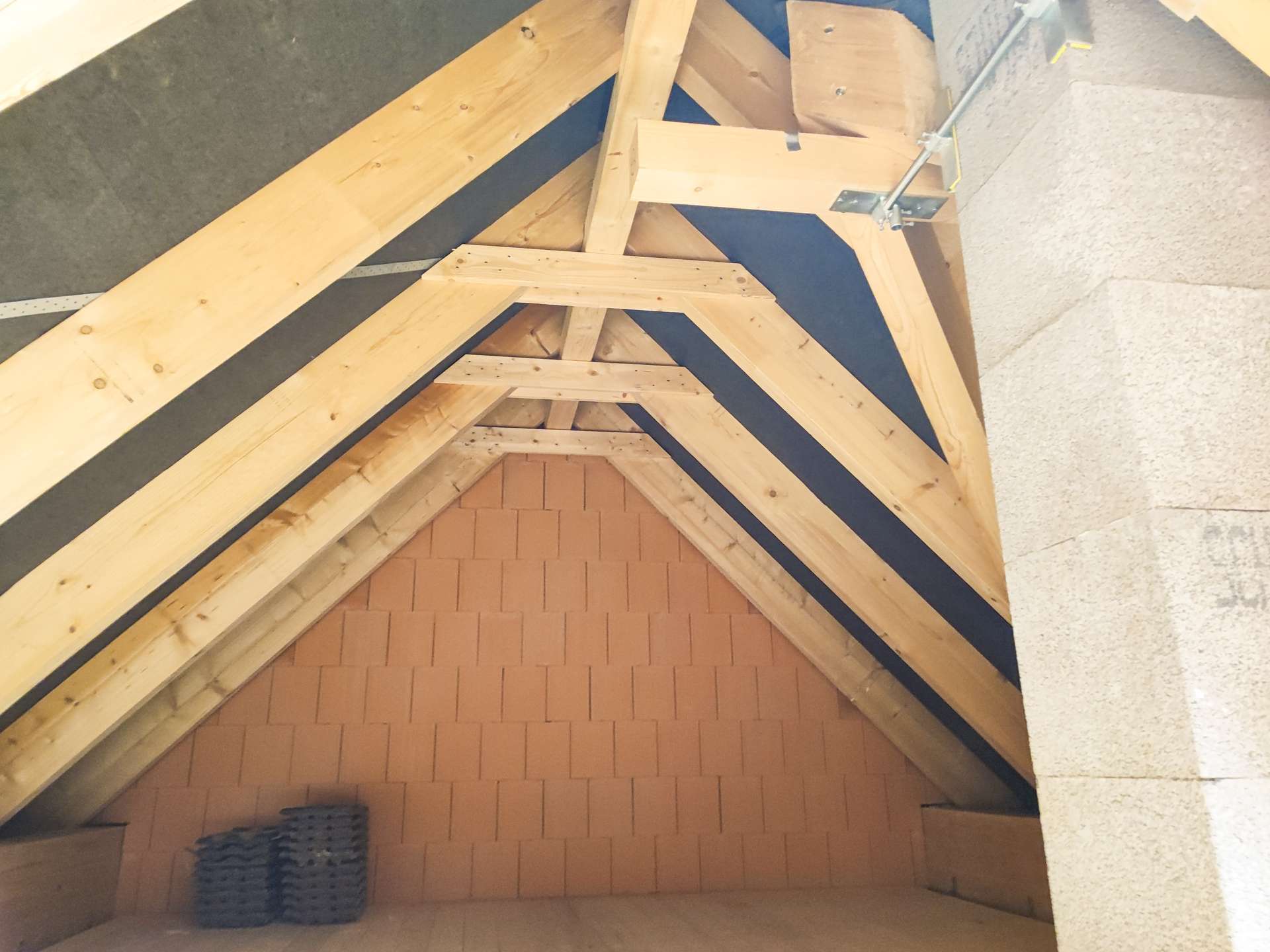 Unfinished attic with exposed wooden roof trusses, brick back wall at the end, and a small stack of roofing tiles on the floor.