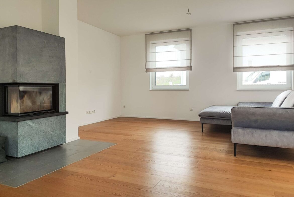 Modern living room with a gray stone fireplace on the left, hardwood floors, and a gray sectional sofa near two large windows with light-filtering blinds.