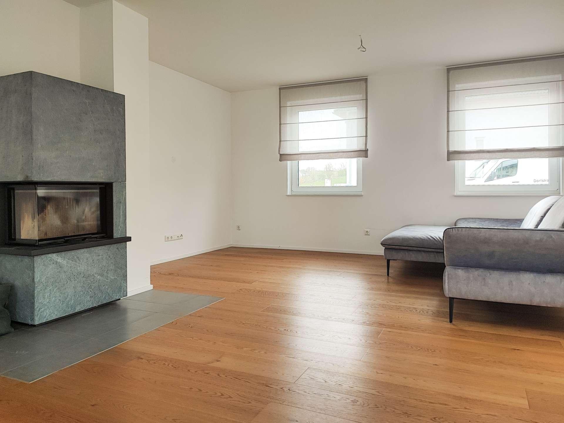 Modern living room with a gray stone fireplace on the left, hardwood floors, and a gray sectional sofa near two large windows with light-filtering blinds.