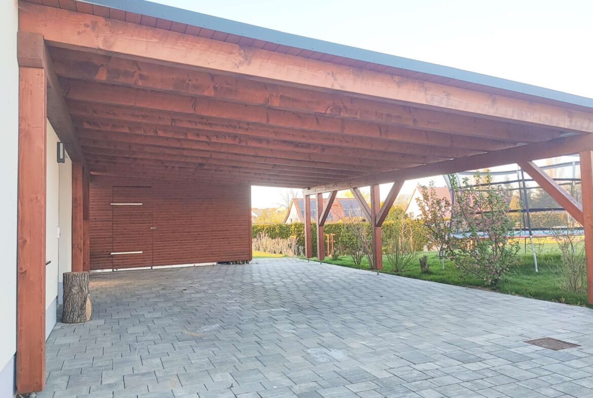 Wooden carport with large beams over a paved driveway; garden area and houses visible in the background.