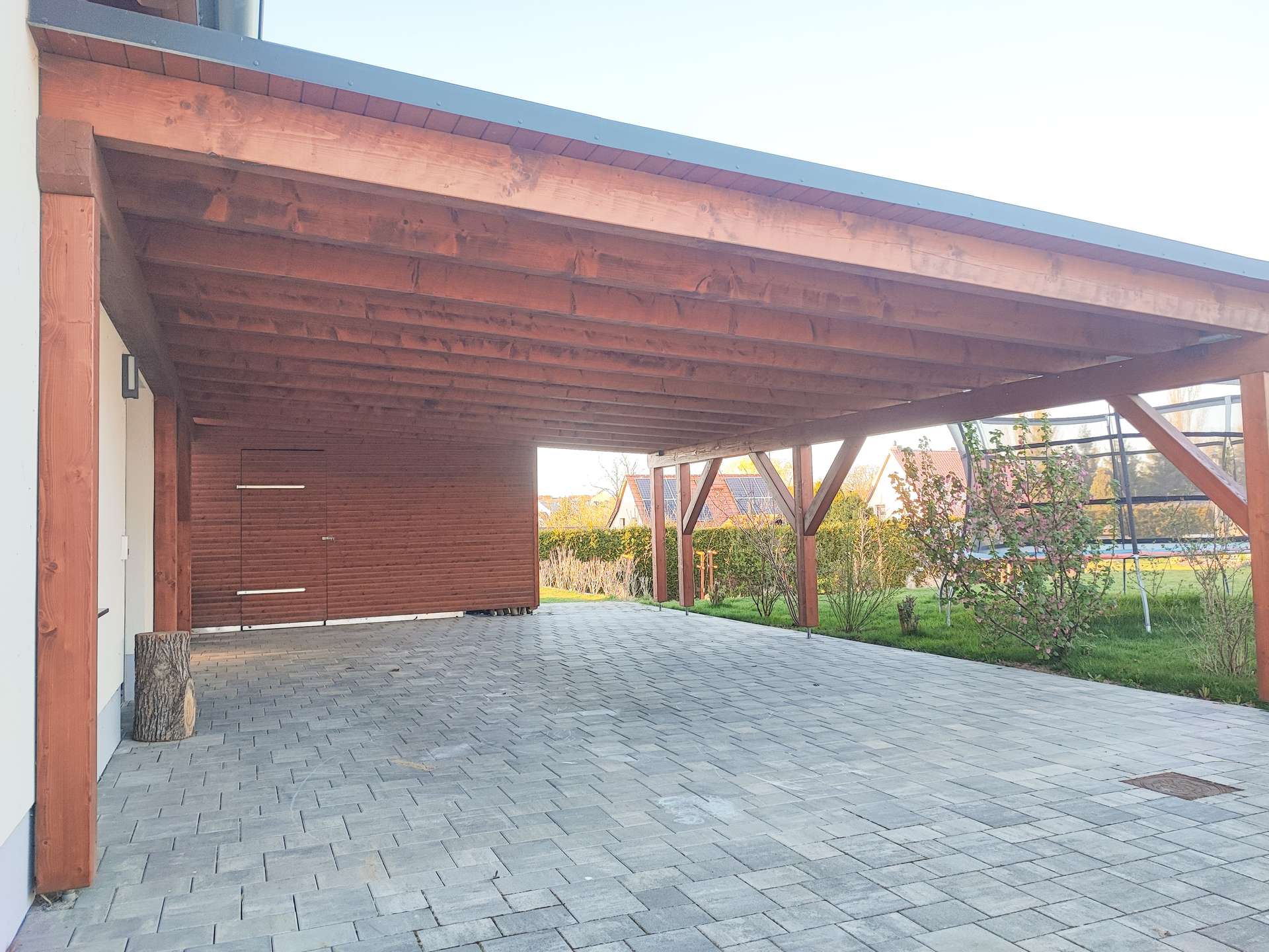 Wooden carport with large beams over a paved driveway; garden area and houses visible in the background.