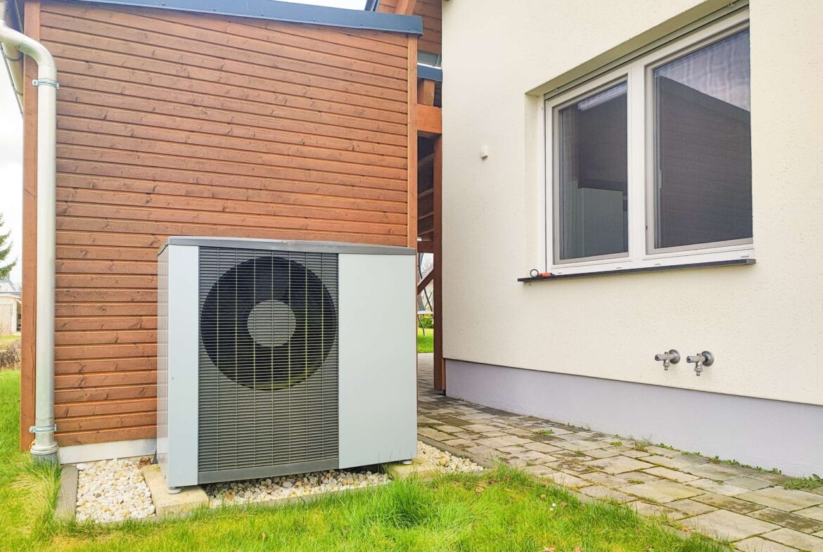 Outdoor air conditioning compressor unit next to a beige house with a wooden wall, window, and gravel bed along the foundation.