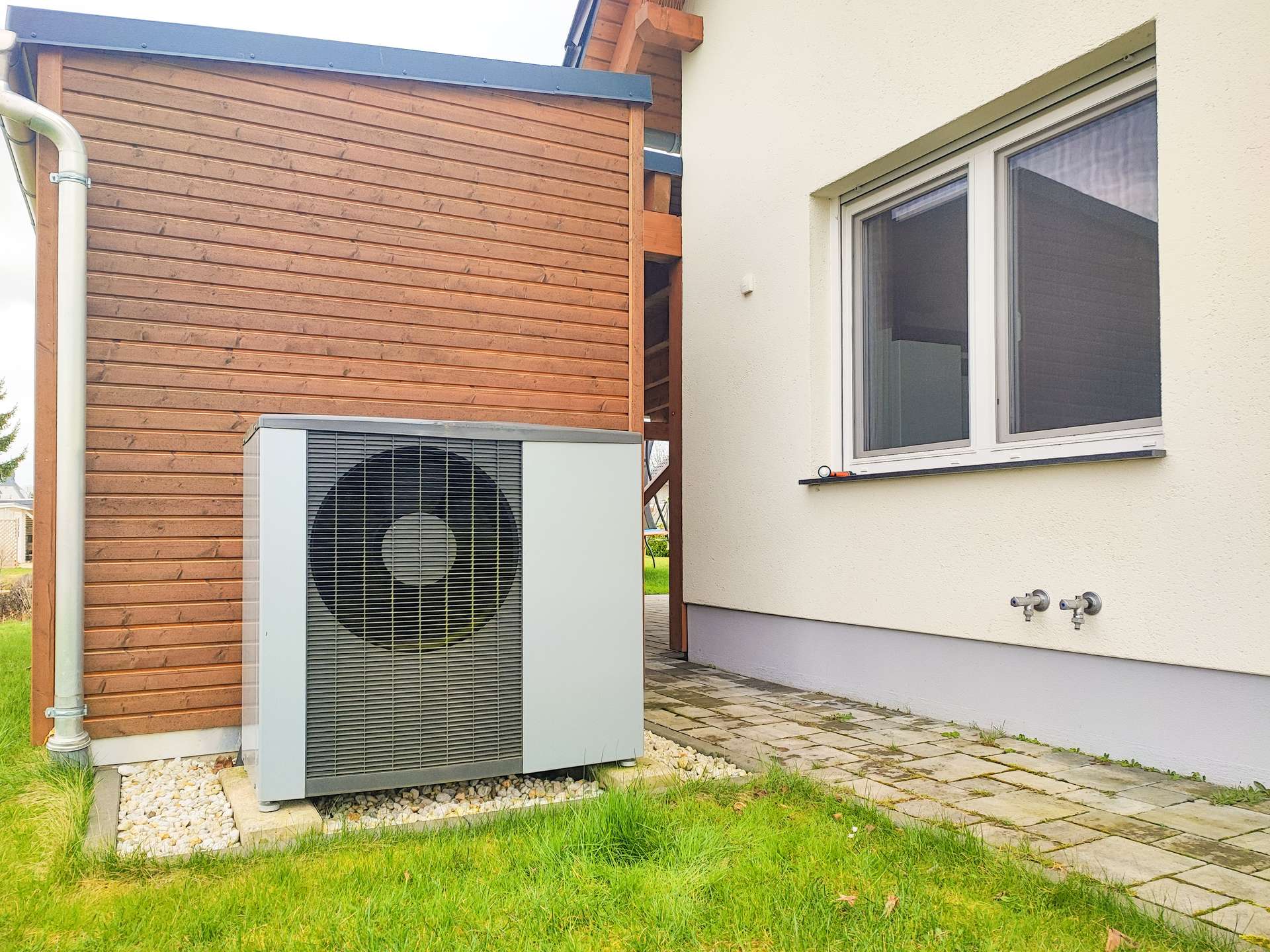 Outdoor air conditioning compressor unit next to a beige house with a wooden wall, window, and gravel bed along the foundation.